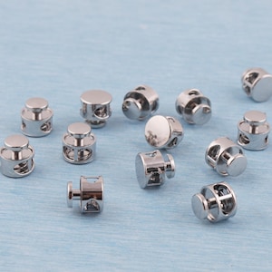 May include: A collection of shiny, silver cord locks. These small, cylindrical fasteners have a button-like top and a hollow, cage-like body with holes. They are designed to secure cords and drawstrings. The background is a light blue.
