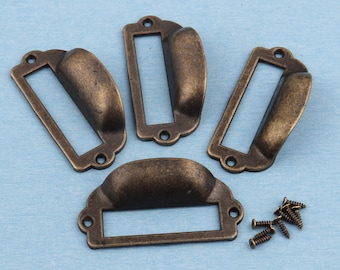 May include: Four antique brass drawer pulls with a rectangular label holder. The pulls have a curved top and a flat bottom. The label holders are rectangular and have a raised edge. The pulls are arranged in a semi-circle with the label holders facing outward.