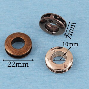 May include: Three circular metal grommets in bronze, gunmetal, and silver tones. The bronze grommet has a 22mm diameter. The gunmetal grommet has a 7mm height. The silver grommet has a 10mm inner diameter. Each piece features a central hole and a smooth, rounded outer edge.