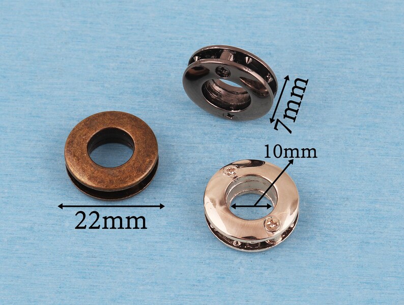 May include: Three circular metal grommets in bronze, gunmetal, and silver tones. The bronze grommet has a 22mm diameter. The gunmetal grommet has a 7mm height. The silver grommet has a 10mm inner diameter. Each piece features a central hole and a smooth, rounded outer edge.