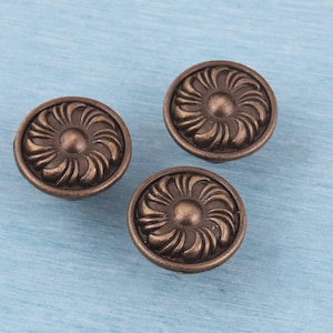 May include: Three bronze-colored cabinet knobs with a floral design. Each knob is round with a raised, detailed pattern resembling a stylized flower. The knobs are set against a light blue background.