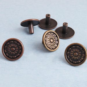 May include: Bronze-colored cabinet knobs with a floral design. The knobs have a round face with a raised flower pattern and a stem for mounting. The knobs are made of metal and are arranged on a light blue surface.