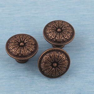 May include: Three bronze-colored cabinet knobs with a floral design. Each knob has a rounded top with a detailed pattern and a short stem. The knobs are arranged on a light blue surface.