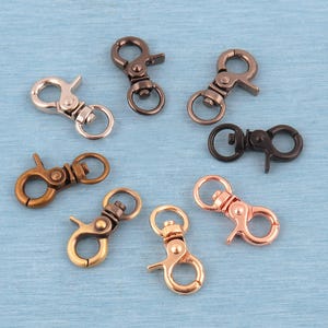 May include: A collection of seven metal lobster claw clasps arranged in a circular pattern. The clasps feature various metallic finishes including silver, antique bronze, copper, rose gold, gunmetal, and gold tones, each with a swivel base.