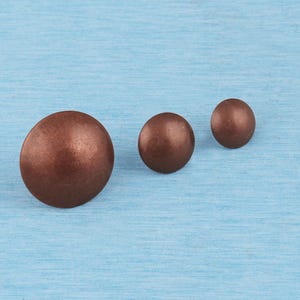 May include: Three round, dome-shaped metal upholstery tacks with a smooth, matte copper-toned finish. The tacks are arranged in a line, decreasing in size from left to right, featuring a solid, circular head design with a metallic surface texture.