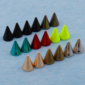 May include: An assortment of cone-shaped metal studs in a range of colours, such as black, teal, red, yellow-green, gold, and silver. The studs are arranged in rows on a light blue surface, highlighting their metallic sheen and pointed design.