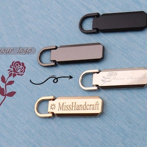 Puede incluir: Tiradores de cremallera metálicos en negro, gris plomo y dorado. Los tiradores rectangulares tienen un lazo para sujetar. Algunos presentan texto grabado y diseños florales.