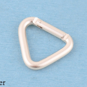May include: A silver-colored metal carabiner in a triangular shape. The hardware features a spring-loaded gate mechanism that allows for opening and closing. The finish is matte and smooth, with the word Silver printed in the bottom left corner.