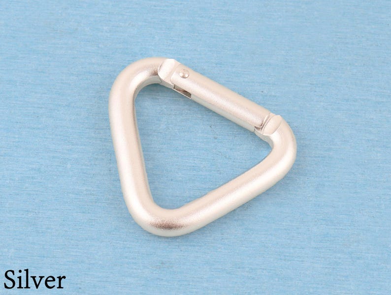 May include: A silver-colored metal carabiner in a triangular shape. The hardware features a spring-loaded gate mechanism that allows for opening and closing. The finish is matte and smooth, with the word Silver printed in the bottom left corner.