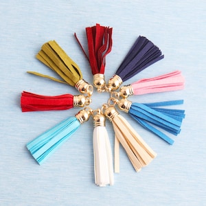 Puede incluir: Una colección de borlas coloridas en varios tonos, incluyendo rojo, azul y amarillo, dispuestas en un patrón circular. Cada borla tiene una tapa dorada. Las borlas están hechas de un material suave similar al ante.