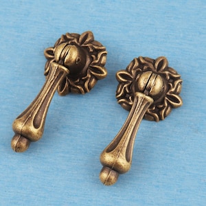 May include: Two antique brass-toned metal drawer pulls featuring a drop-style handle attached to a circular backplate with a raised floral petal design. The hardware has a weathered finish with dark recessed areas and lighter highlights.