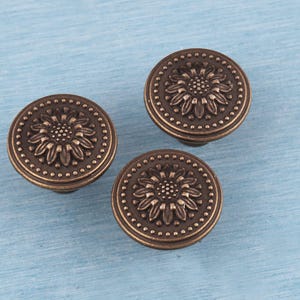 May include: Three bronze-colored cabinet knobs with a sunflower design. Each knob features a detailed floral center surrounded by a dotted border. The knobs are round and have a vintage aesthetic, suitable for furniture restoration or decorative projects.