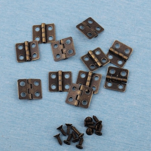 May include: A collection of small, antique bronze-colored hinges with matching screws. The hinges are square with rounded corners and four screw holes. The hardware is scattered on a light blue surface.