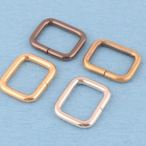 Könnte beinhalten: Vier rechteckige Metallringe, die in einer Gruppe angeordnet sind. Jeder Ring weist ein unterschiedliches metallisches Finish auf, darunter dunkle Bronze, mattes Gold, poliertes Gold und ein Roségold-Ton. Die Ringe haben abgerundete Ecken.