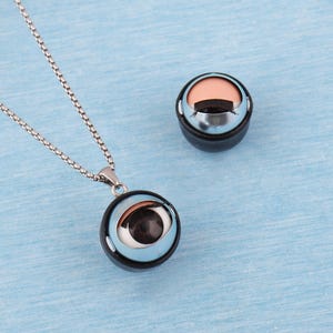 Puede incluir: Un collar con cadena plateada y colgante redondo con diseño de ojo. El colgante tiene un borde negro, un anillo interior azul claro y un ojo detallado. Un objeto a juego con forma de ojo se encuentra junto al collar.