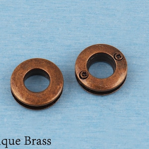 May include: Two circular antique brass grommets with a distressed metallic finish. Each grommet features a central opening and a flat outer ring with two small, dark, recessed screw-like details visible on the surface of the right-hand piece.