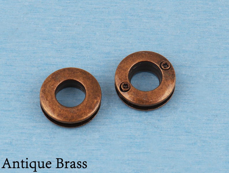 May include: Two circular antique brass grommets with a distressed metallic finish. Each grommet features a central opening and a flat outer ring with two small, dark, recessed screw-like details visible on the surface of the right-hand piece.