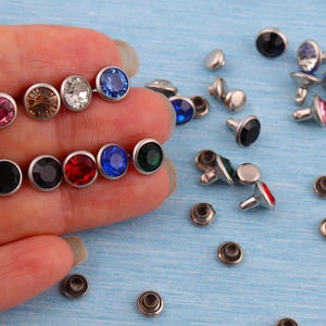 May include: A collection of colorful rhinestone rivets with silver-colored metal settings. The rivets feature various jewel tones, including pink, brown, clear, blue, red, and green. Some rivets are scattered on a light blue surface.