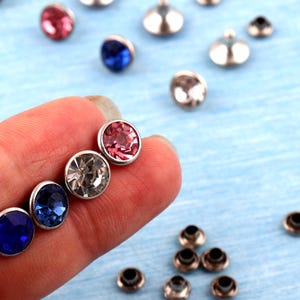 Puede incluir: Primer plano de varios remaches de pedrería de colores con bases de metal plateado. Los remaches son en tonos rosa, azul y transparente. Algunos remaches se sostienen en una mano, mientras que otros están dispersos sobre una superficie azul claro.