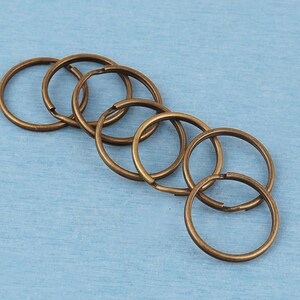 May include: A collection of six circular metal split rings with an antique bronze finish. Each ring features a double-loop coil design, commonly used for attaching keys or charms. The rings are arranged in a slightly overlapping diagonal line.