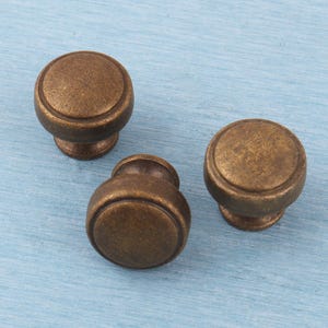 May include: Three antique bronze cabinet knobs with a round, stepped design. The knobs are made of metal and have a warm, aged finish. They are arranged on a light blue surface.