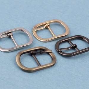 May include: Four metal belt buckles in different finishes: silver, gold, bronze, and black. Each buckle has a rectangular shape with a rounded edge and a single prong.