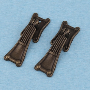 May include: Two antique bronze-toned metal drop pulls featuring a vertical fluted design on the upper section and a smooth, flared base. Each handle is attached to a decorative mounting plate with a hinged mechanism at the top for movement.