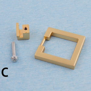 Op de afbeelding: Een set goudkleurige kastbeslagonderdelen, waaronder een vierkante handgreep, een montageblok en een schroef. De handgreep heeft een vierkante vorm met een rechthoekige opening. De letter "C" is links onderaan in het zwart gedrukt.