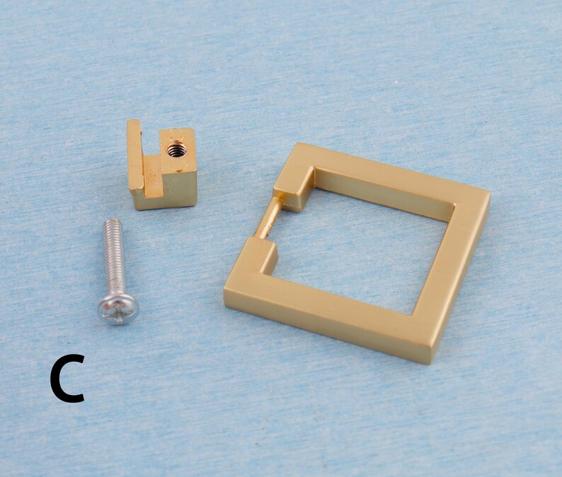 Op de afbeelding: Een set goudkleurige kastbeslagonderdelen, waaronder een vierkante handgreep, een montageblok en een schroef. De handgreep heeft een vierkante vorm met een rechthoekige opening. De letter "C" is links onderaan in het zwart gedrukt.
