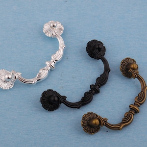 May include: Three decorative drawer pulls in silver, black, and antique brass. Each pull features a floral design on the ends and a curved handle with ornate detailing. The pulls are set against a light blue background.