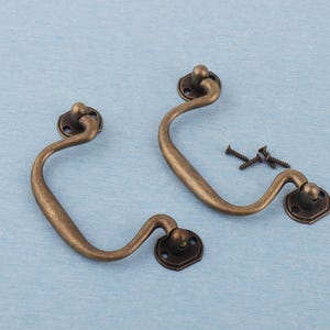 May include: Two antique bronze drawer pulls with screws. Each pull features a curved handle and decorative base plates. The hardware is set against a light blue background, suggesting a vintage or rustic aesthetic.