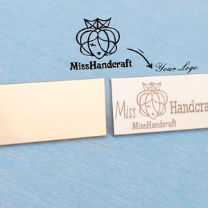 May include: Two rectangular metallic tags with a polished finish. One tag is blank, while the other features an engraved logo of a stylized crown and face above the text Miss Handcraft. The design includes a graphic showing the logo and text Your Logo.
