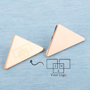 May include: Two rose gold triangle-shaped objects with rounded corners. One has a logo with the letters "J", "N", and "&" inside overlapping squares. The other object is blank. The image includes the text "Your Logo".