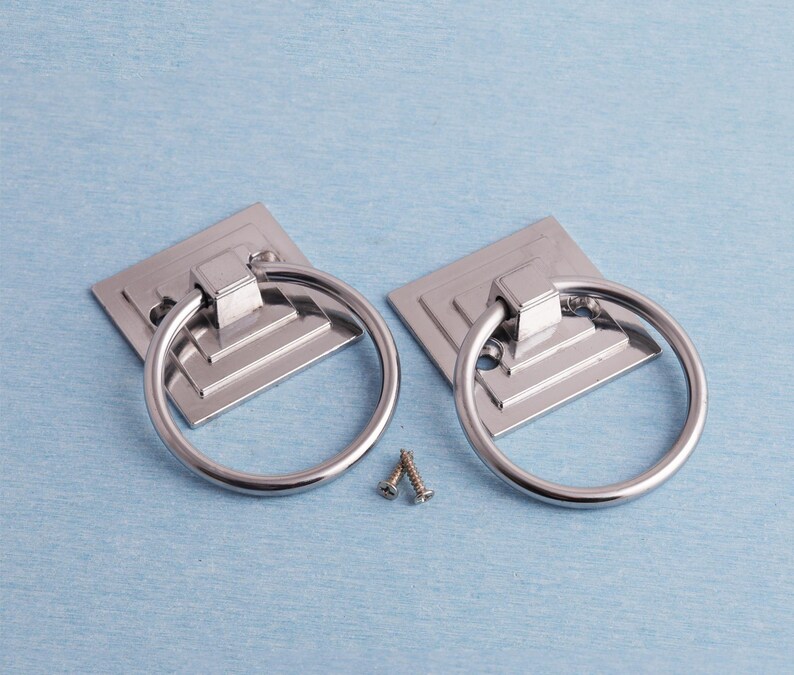 K&ouml;nnte beinhalten: Zwei silberfarbene Schrankgriffe mit quadratischen Grundplatten und runden Griffen. Zwei kleine Schrauben sind ebenfalls zu sehen. Sie liegen auf einem hellblauen Hintergrund. Geeignet f&uuml;r M&ouml;bel.