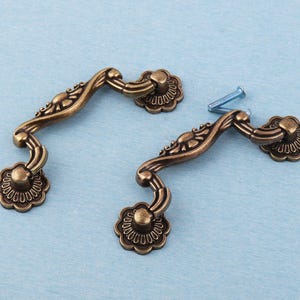May include: Two antique bronze drawer pulls with a floral design. Each pull has a curved handle and decorative floral end pieces. Two blue screws are also visible, suggesting installation hardware.