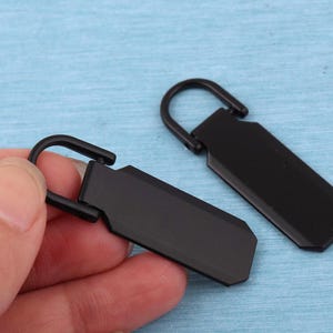 Peut inclure: Deux tirettes de fermeture &eacute;clair en m&eacute;tal noir mat, chacune dot&eacute;e d'un corps rectangulaire aux coins biseaut&eacute;s et d'une boucle articul&eacute;e arrondie sur le dessus pour la fixation. Le design est &eacute;pur&eacute; et uniforme avec une finition sombre.