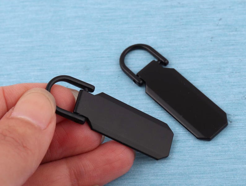 Peut inclure: Deux tirettes de fermeture &eacute;clair en m&eacute;tal noir mat, chacune dot&eacute;e d'un corps rectangulaire aux coins biseaut&eacute;s et d'une boucle articul&eacute;e arrondie sur le dessus pour la fixation. Le design est &eacute;pur&eacute; et uniforme avec une finition sombre.