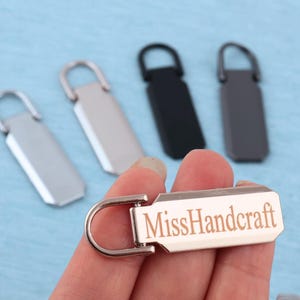 Peut inclure: Un ensemble de tirettes de fermeture &eacute;clair rectangulaires en m&eacute;tal avec une boucle sup&eacute;rieure arrondie. Une tirette est tenue au premier plan, portant le texte grav&eacute; MissHandcraft. L'ensemble comprend des finitions argent, noir et gris fonc&eacute;.