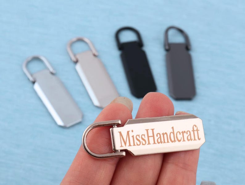 Peut inclure: Un ensemble de tirettes de fermeture &eacute;clair rectangulaires en m&eacute;tal avec une boucle sup&eacute;rieure arrondie. Une tirette est tenue au premier plan, portant le texte grav&eacute; MissHandcraft. L'ensemble comprend des finitions argent, noir et gris fonc&eacute;.