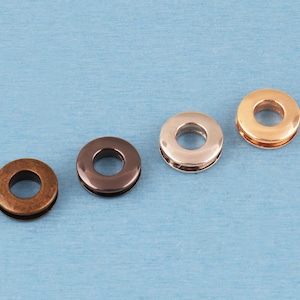 May include: Four circular metal grommets arranged in a diagonal line. The grommets feature distinct finishes including a matte antique bronze, a polished dark gunmetal, a smooth rose gold, and a bright polished gold tone, each with a center hole.