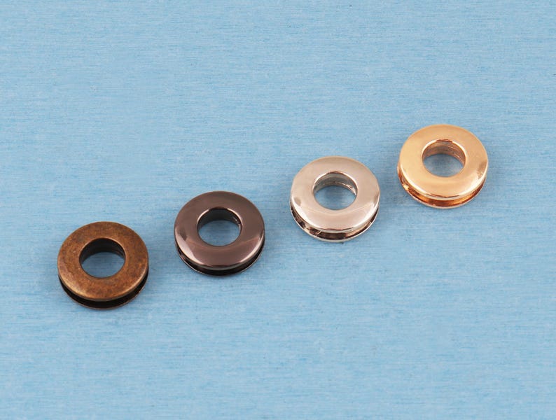 May include: Four circular metal grommets arranged in a diagonal line. The grommets feature distinct finishes including a matte antique bronze, a polished dark gunmetal, a smooth rose gold, and a bright polished gold tone, each with a center hole.