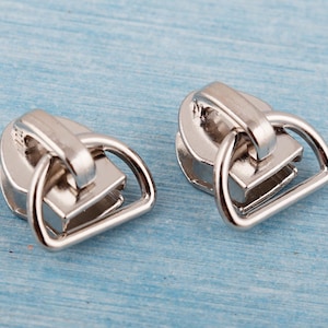 May include: Two silver-colored zipper pulls with a rounded top and a square base. Each pull has a small, attached ring. The pulls are displayed on a light blue, textured surface.