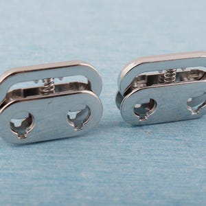 May include: Two silver metal clasps with a spring mechanism. Each clasp has an oval shape with two keyhole-shaped openings and a curved top. The clasps are designed to connect two pieces together. The background is a light blue.
