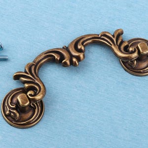 May include: A bronze-toned metal drawer pull with an ornate, curved design featuring scrollwork and leaf-like details. The handle is attached to two circular mounting plates and is accompanied by two small silver-colored metal screws.