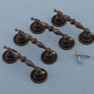 May include: Four antique bronze cabinet pulls with decorative detailing. Each pull features a round backplate with a beaded edge and a curved handle. Two silver screws are also included.
