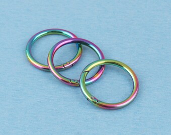 May include: Three circular metal spring rings featuring a vibrant iridescent rainbow finish. Each ring has a hinged opening mechanism with a small visible spring-loaded gate, allowing the circular loop to open and close securely for attachment.