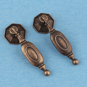 Puede incluir: Dos tiradores de cajón de metal en tono bronce antiguo con una placa octogonal de diseño escalonado y un asa ovalada alargada colgante con punta redondeada. Los herrajes tienen un acabado texturizado y desgastado con tonos oscuros.