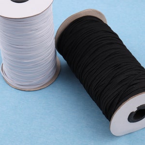 May include: Two spools of elastic cord, one white and one black, are wound around cardboard spools. The spools are lying on a light blue surface.