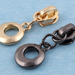 May include: Two zipper pulls, one gold and one black, with a round ring at the bottom. The pulls are made of metal and have a shiny finish.