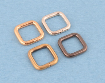 May include: Four square metal rings in different metallic finishes, including rose gold, silver, matte gold, and dark bronze. Each ring features a smooth, rounded profile with a visible seam where the ends meet, arranged in a cluster.